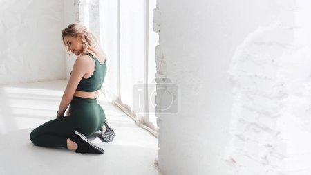 Photo for Blonde fitness woman posing on the floor in studio and stretching while looking down over white background - Royalty Free Image