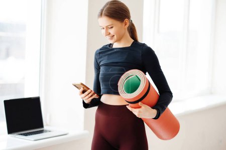 Photo for Beautiful young blonde in sport clothes standing near window with laptop, holding fitness rug in hand and reading message on smartphone. Smiling lady finishing workout in bright gym. - Royalty Free Image
