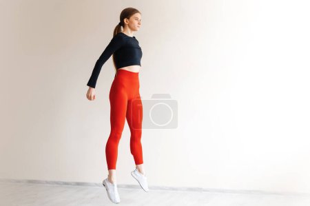 Photo for Active healthy woman doing cardio exercises and jumping in front of windows of white studio. Young energetic female dressed in tight sports clothes training legs muscles. - Royalty Free Image