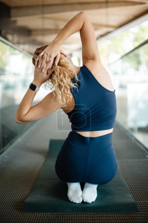 Foto de Vista trasera de la atleta rubia en ropa deportiva ejercitando y estirando el cuerpo en la calle mientras está sentada en la esterilla de yoga. Concepto deportivo - Imagen libre de derechos