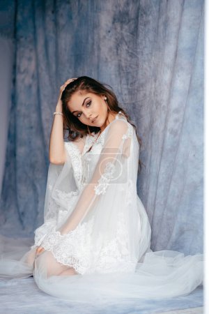 Téléchargez les photos : Full length view of the young brunette bride in white luxury wedding dress looking at the camera at loft studio interior with blue fabric background - en image libre de droit