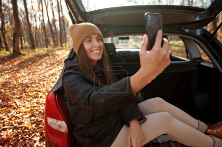 Photo for Happy stylish hipster woman in beige hat holding phone and doing selfie. Woman sitting in car trunk at sunset light. Travel and autumn road trip. Young female with smartphone traveling - Royalty Free Image