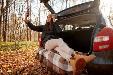 Téléchargez les photos : Vue en angle bas de la femme positive faisant du selfie sur le coffre de la voiture pendant le voyage de l'automne. Voyage en forêt d'automne en voiture concept - en image libre de droit