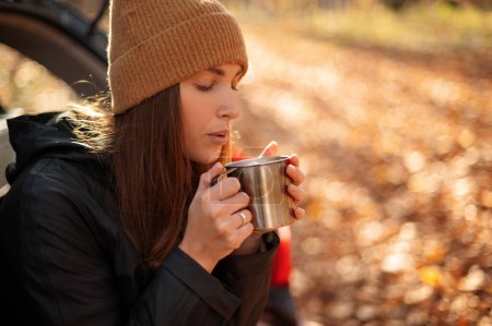 Photo for Focused girl sitting in open car trunk and blowing on hot tea during autumn day. Adventure road trip and traveller lifestyle concept - Royalty Free Image
