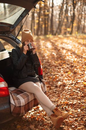 Téléchargez les photos : Vue verticale de la fille caucasienne assise dans le coffre de la voiture ouverte et dégustant du thé chaud pendant la journée d'automne. Aventure road trip et mode de vie du voyageur concept - en image libre de droit