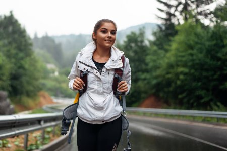 Photo for Happy young woman with backpack walking on wet road and enjoying beautiful view on mountains. Foggy and rainy weather outdoors. Active time spending. - Royalty Free Image