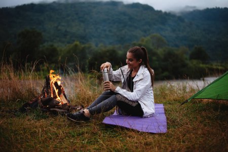 Foto de Feliz joven mujer vertiendo té de la botella de termo mientras está sentado cerca de la quema de fuego al aire libre. Viajera disfrutando de bebidas calientes durante el campamento en la naturaleza. - Imagen libre de derechos
