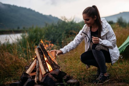 Téléchargez les photos : Positive young woman preparing bonfire. Joyful woman enjoying of the camping at the nature near her tent. Girl alone at the forest - en image libre de droit
