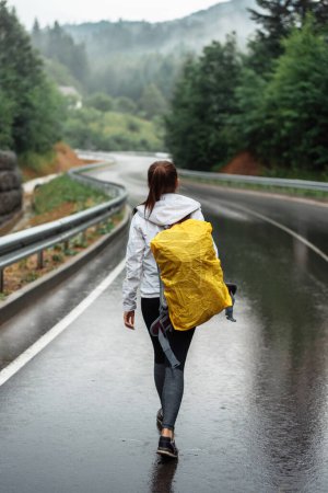 Téléchargez les photos : Back view of the young woman going with yellow backpack in nature. Happy girl is trekking on forest road with camping stuff. Travel freedom lifestyle - en image libre de droit