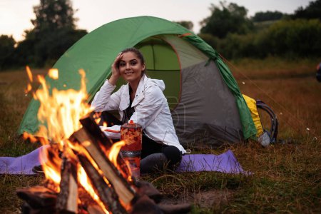 Foto de Happy backpacker woman sitting in entrance of tourist tent at campfire and looking away near forest under sky. Camping at the forest concept - Imagen libre de derechos