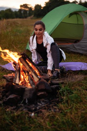 Photo for Beautiful female traveler smiling while looking at the bonfire and resting on grass near bonfire. Tourism concept. Stock photo - Royalty Free Image