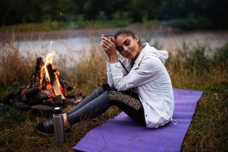 Téléchargez les photos : Travel, tourism, camping. Young calm woman tourist at the beautiful nature landscape sitting near the tent by the fire. Girl with drink in mug in her hands - en image libre de droit