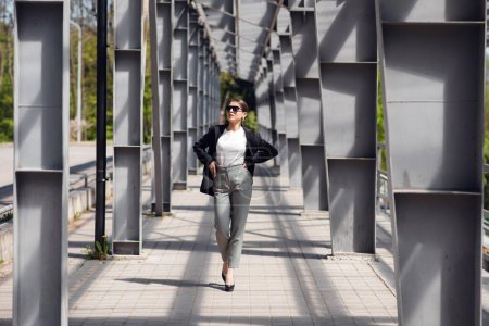 Téléchargez les photos : Femme d'affaires élégante marchant à travers le pont avec une émotion confiante pendant sa pause dîner. Concept de style de vie - en image libre de droit