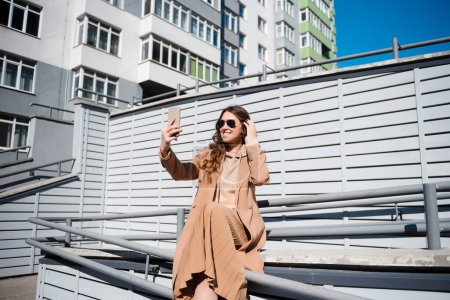 Photo for Young smiling brunette woman in coat and sunglasses makes selfie outdoors on a sunny day, against striped wall background. - Royalty Free Image
