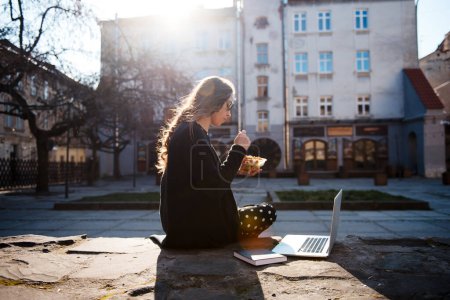 Téléchargez les photos : Jeune femme assise dehors, mangeant de la salade de légumes et travaillant à l'ordinateur portable sur fond de ville urbaine. Printemps journée ensoleillée. Travail à l'extérieur, déjeuner - en image libre de droit