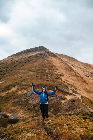 Foto de Mujer alegre caucásica en chaqueta azul con modernos bastones de trekking en las manos de pie entre las altas montañas de la cima. HIking, actividad de ocio y concepto de personas. - Imagen libre de derechos