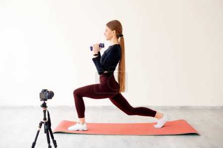 Photo for Professional personal trainer recording video on camera during physical activity for own followers. Flexible fitness woman doing leg workout exercises and practicing lunges with dumbbells on mat. - Royalty Free Image