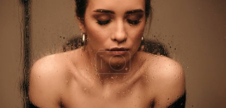 Téléchargez les photos : Vue à travers verre mouillé sur belle jeune femme aux cheveux bruns posant sensuellement en studio. Modèle féminin caucasien portant un pull noir avec épaule nue et boucles d'oreilles rondes dorées - en image libre de droit