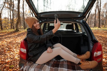 Foto de Feliz mujer hipster elegante en sombrero beige sosteniendo el teléfono y haciendo selfie. Mujer sentada en el maletero al atardecer. Viaje y viaje de otoño por carretera. Mujer joven con smartphone viajando - Imagen libre de derechos