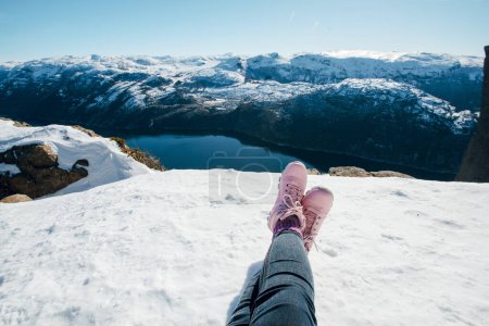 Foto de Patas de mujer con botas rosas. Mujer sentada en la nieve. Paisaje escénico del canal fluvial, mar entre ribera rocosa con nieve. Vista superior de la roca del púlpito, Preikestolen. Lysefjord, Noruega - Imagen libre de derechos