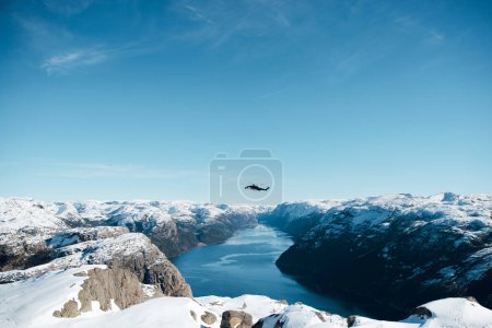Foto de Helicóptero volando sobre el lago, el canal del río, el mar entre la costa rocosa con nieve. Vista superior de la roca del púlpito, Preikestolen. Montañas noruegas. Lysefjord, Noruega - Imagen libre de derechos