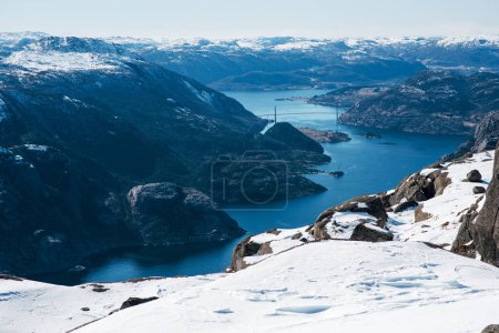 Foto de Maravilloso paisaje de canal de lago, mar entre costa rocosa con nieve. Puente en el fondo. Vista superior de la roca del púlpito, Preikestolen. Montañas noruegas. Lysefjord, Noruega - Imagen libre de derechos