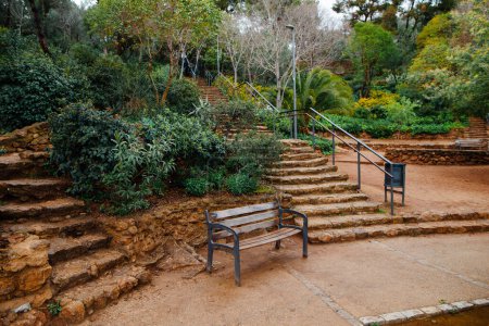 Foto de Un banco de madera en el callejón entre el jardín verde en el parque de primavera de Guell en España. Sendero y escalones de piedra que conducen al impresionante mirador de Barcelona. - Imagen libre de derechos