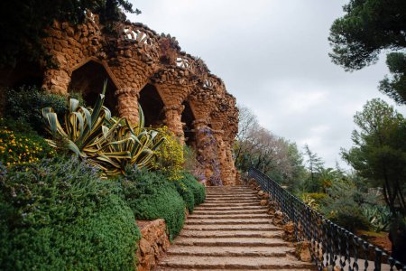Foto de Escalera a hermosas columnas en el parque Gaudí de Barcelona. Arcos de piedra cerca de exóticos arbustos verdes y plantas de flores en España. - Imagen libre de derechos
