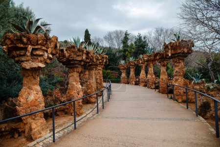Foto de Walking road through stone columns decorated with exotic bushes on top at Guell park in Barcelona. Concept of tourism and old architecture. - Imagen libre de derechos