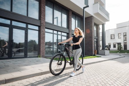 Photo for Caucasian female athlete on sport clothes walking on street with black bike in hands. Young female blonde preparing for morning cycling on fresh air. - Royalty Free Image