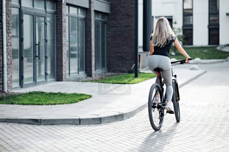 Téléchargez les photos : Vue arrière de la jeune femme vêtue de vêtements de sport à vélo sur la rue de la ville. Blonde caucasienne active passant le matin à faire du vélo. Mode de vie sain concept. - en image libre de droit