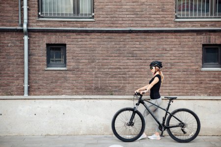 Foto de Vista lateral de ciclista femenina caucásica en ropa deportiva, casco y gafas de pie con bicicleta negra en la calle de la ciudad. Relajación después del entrenamiento. - Imagen libre de derechos