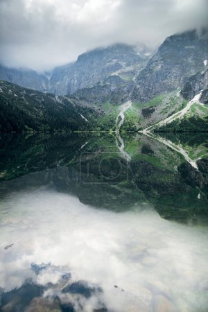 Téléchargez les photos : Belle vue sur les montagnes brumeuses couvertes de nuages sombres et de forêt verte avec un reflet dans un lac. Rivage pierreux. Morskie Oko. Oeil marin. High Tatras, Zakopane, Pologne - en image libre de droit
