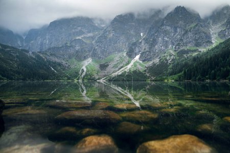 Téléchargez les photos : Belle vue sur les montagnes brumeuses couvertes de nuages sombres et de forêt verte avec un reflet dans un lac. Rivage pierreux. Morskie Oko. Oeil marin. High Tatras, Zakopane, Pologne - en image libre de droit