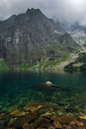 Photo for Scenic view of foggy mountains cover by dark clouds and green forest with a reflection in a lake. Stony shore. Morskie Oko. Marine Eye. High Tatras, Zakopane, Poland - Royalty Free Image