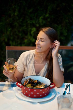 Photo for Beautiful caucasian woman sitting at dining table on fresh air with large bowl of seafood and glass of wine. Charming smiling lady keeping eyes closed and adjusting hair with fingers. - Royalty Free Image