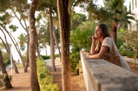Photo for Smiling adorable lady looking at stunning scenery of Brac Island while leaning on balusters. Pretty brunette dressed in casual clothes and holding beach bag exploring sightseeing of Croatia. - Royalty Free Image