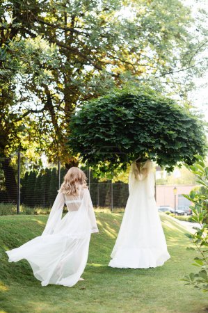 Téléchargez les photos : Vue arrière de la jeune femme avec une coiffure blonde allant à sa belle robe de mariée qui pendent à l'arbre. Préparation matinale de charmante mariée. - en image libre de droit
