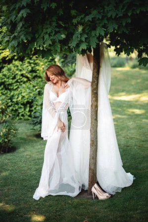 Photo for Full length portrait of gorgeous caucasian woman wearing stylish lingerie and dressing gown standing near wedding dress that hanging on tree. Bride getting ready for event outdoors. - Royalty Free Image
