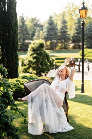 Téléchargez les photos : Mariée caucasienne joyeuse avec maquillage de mariage et coiffure reposant sur une chaise à l'extérieur pendant la matinée. Profiter de chaque instant. Des émotions humaines positives. - en image libre de droit
