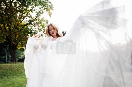 Photo for Attractive woman with trendy hairstyle and makeup holding on hanger her wedding dress while standing outdoors. Concept of bride's morning. - Royalty Free Image