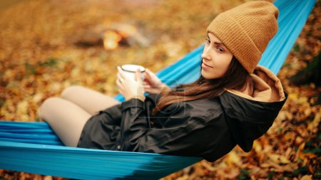 Téléchargez les photos : Vue latérale de charmante femme caucasienne en vêtements chauds et chapeau assis dans un hamac bleu avec tasse de boisson chaude. Jolie femme passant du temps libre dans la forêt d'automne. - en image libre de droit