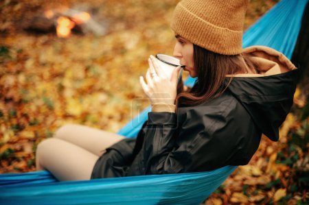 Photo for Close up of relaxed caucasian woman sitting in blue hammock at autumn forest and drinking tea or coffee. Leisure time on fresh air. - Royalty Free Image