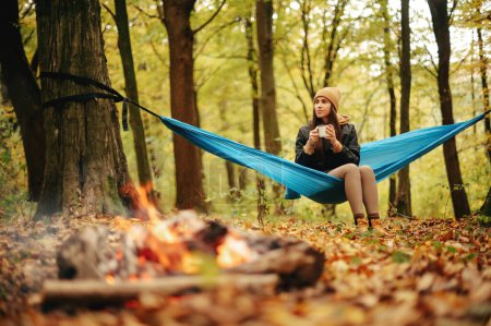 Foto de Encantadora joven en ropa casual caliente beber té caliente y relajarse en hamaca azul entre el hermoso bosque de otoño. Camping en la naturaleza. Concepto de personas y tiempo libre. - Imagen libre de derechos