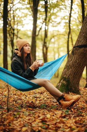 Photo for Pretty caucasian lady in warm clothes and hat drinking hot tea while sitting in hammock among autumn forest. Camping time of female person. - Royalty Free Image