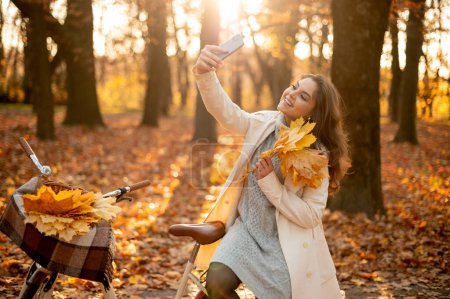 Téléchargez les photos : Femme heureuse assise à son vélo et faisant du selfie dans le parc d'automne. Mode d'automne, mode de vie, détente et concept nature - en image libre de droit
