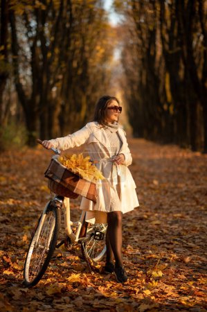 Photo for Vertical view of the young pretty woman posing with vintage white bicycle in autumn park. Lady having fun on orange nature fall background - Royalty Free Image