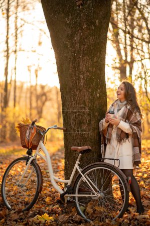 Téléchargez les photos : Vue verticale de la femme souriante se détendre dans la forêt d'automne, tenant une tasse de café, regardant la caméra. Concept de mode de vie actif - en image libre de droit