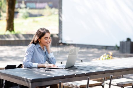 Foto de La sonriente mujer de negocios europea usa computadoras portátiles al aire libre. Hermosa joven se sienta a la mesa en el soleado parque. Estilo de vida femenino moderno. Concepto de trabajo remoto e independiente - Imagen libre de derechos