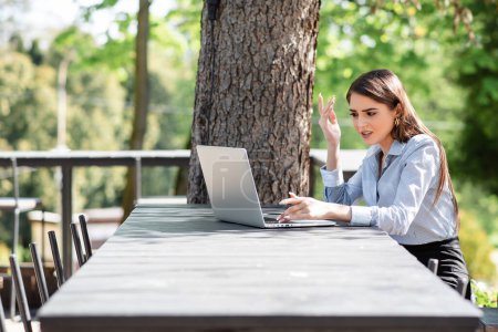 Photo for European dissatisfied businesswoman work on laptop computer outdoors. Beautiful young girl sit at table in sunny park. Modern woman lifestyle. Concept of remote and freelance work - Royalty Free Image